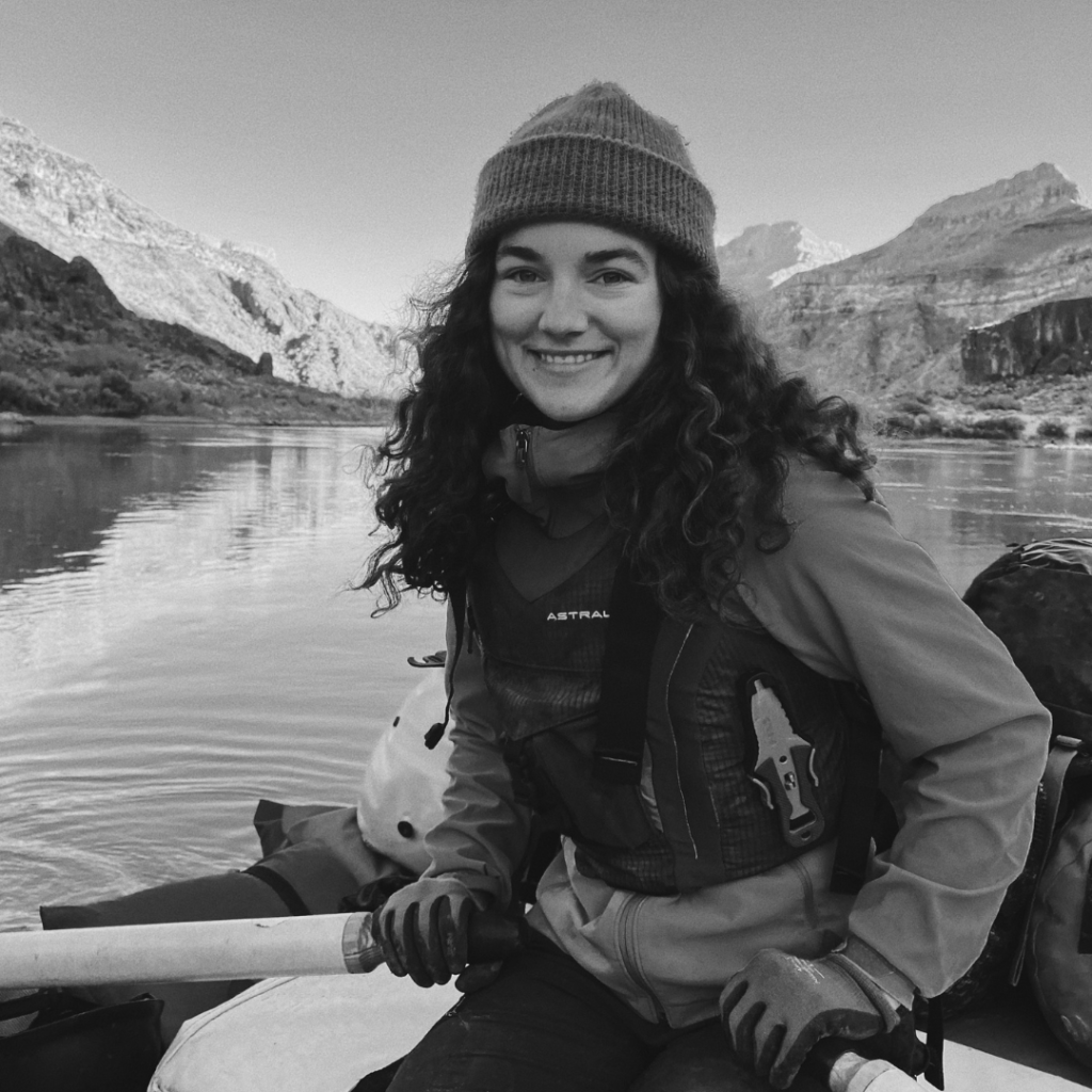 A picture of Sara - Rowing a raft, wearing a beanie with snowy mountains in the background.